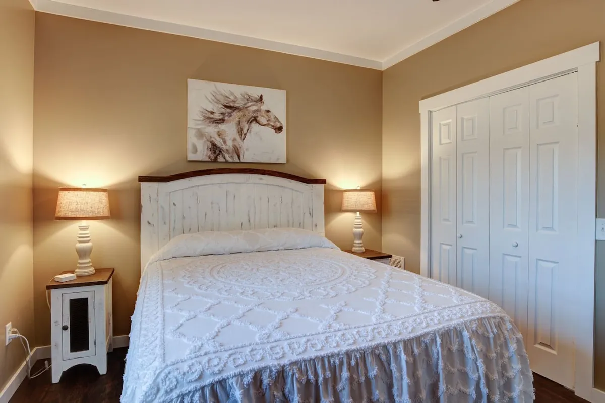 A bright, tranquil bedroom featuring a comfortable, inviting bed flanked by nightstands and softly glowing lamps. Above the bed, a striking horse painting adds a touch of local character. Sunlight pours in, accentuating the room's serene and restful ambiance.