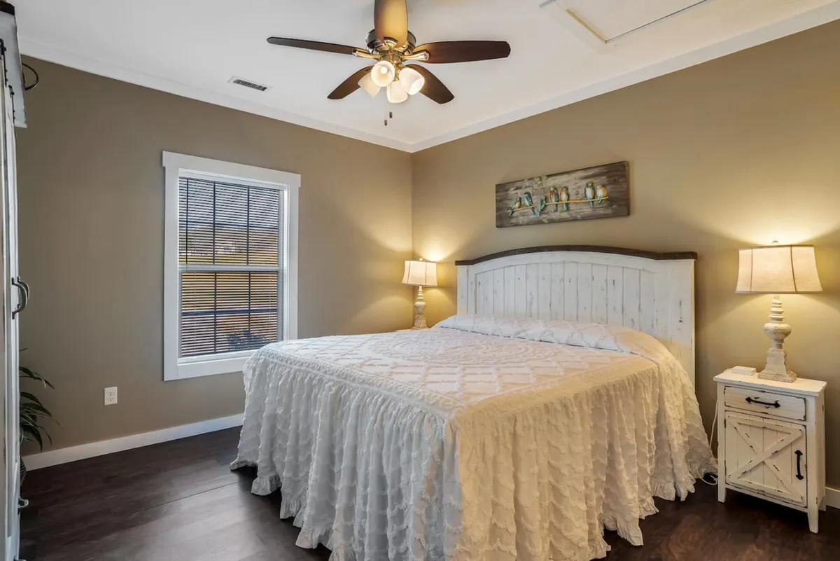 This image captures a serene bedroom with a bed dressed in a white quilted coverlet, a wooden headboard, a nightstand with a lamp, decorative wall art, and a window with blinds. The space is filled with soft natural light, creating a cozy and inviting atmosphere ideal for relaxation or quiet moments.