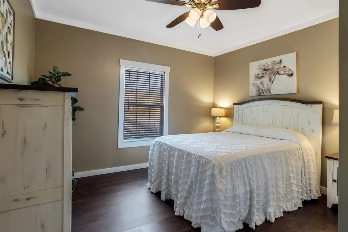 A tranquil bedroom featuring a bed with crisp white linens, a striking horse artwork above the headboard, a decorative plant, and a table lamp. Sunlight filters in, enhancing the restful and fresh feel of the space.
