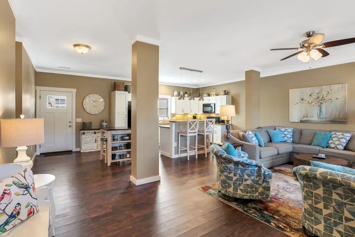 The image showcases a bright, inviting living area with plush, comfortable seating positioned beside a modern, fully equipped kitchen. Tasteful decorative wall art accents the space, which is designed for social gatherings, cozy family meals, and relaxed conversations. The overall ambiance is warm and welcoming, perfect for making memories together.