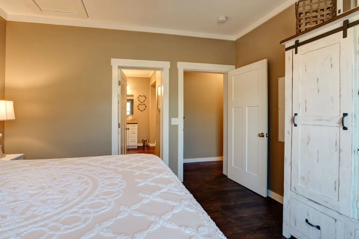 A cozy, light-filled bedroom with intricately patterned bedding, a sturdy wooden cabinet, and a doorway that leads to the bathroom. The space feels serene and restful, with gentle natural light suggesting relaxed mornings and a peaceful retreat.