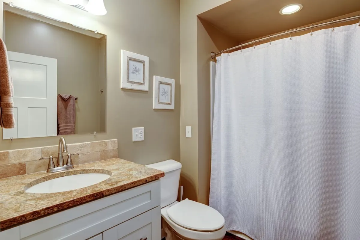 A bright and serene bathroom scene featuring a vanity sink, a soft shower curtain, and framed artwork. Morning sunlight pours through the window, illuminating the calm space and giving a sense of peaceful privacy. The ambiance suggests a quiet moment of relaxation and preparation before venturing out for the day.