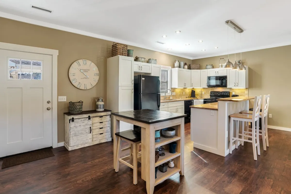 A bright, modern kitchen centered around a spacious island with bar stools, black appliances, and crisp white cabinetry. The kitchen feels inviting and functional, with an open layout that encourages both cooking and socializing. A wall clock adds a practical touch, and the scene suggests activity—perhaps friends baking cookies or preparing brunch together.