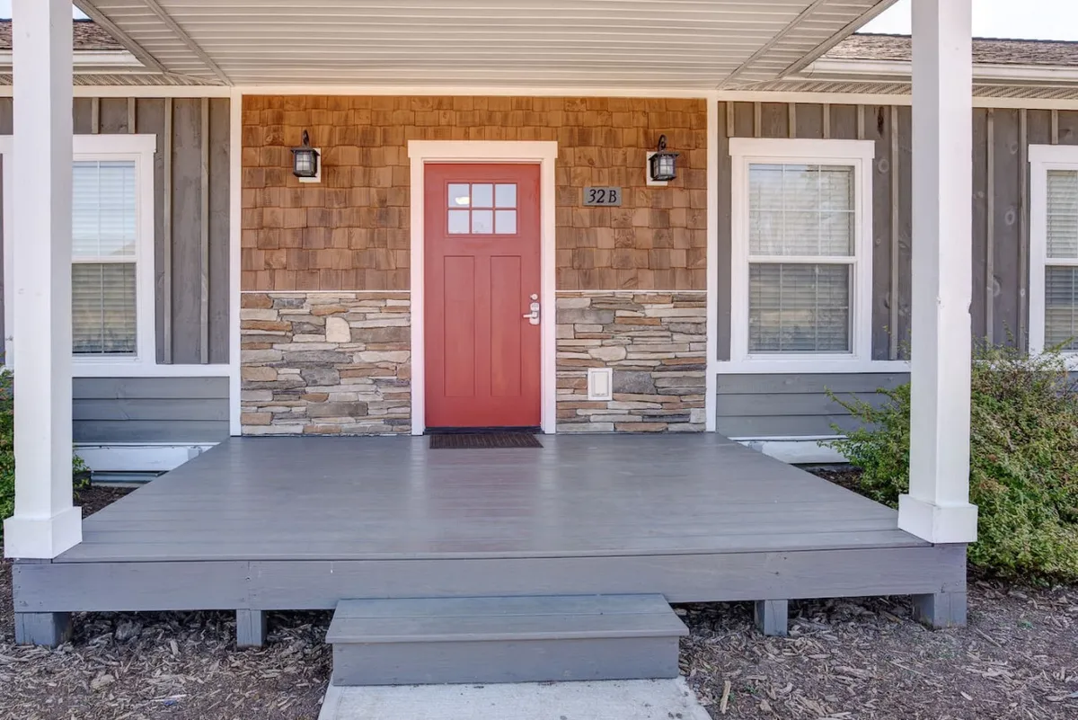 A warm, inviting exterior of the property featuring a striking red front door against a stone facade and a classic wooden porch. The porch is ready to welcome guests, surrounded by simple seasonal decor, and the scene hints at both privacy and a connection to nature.